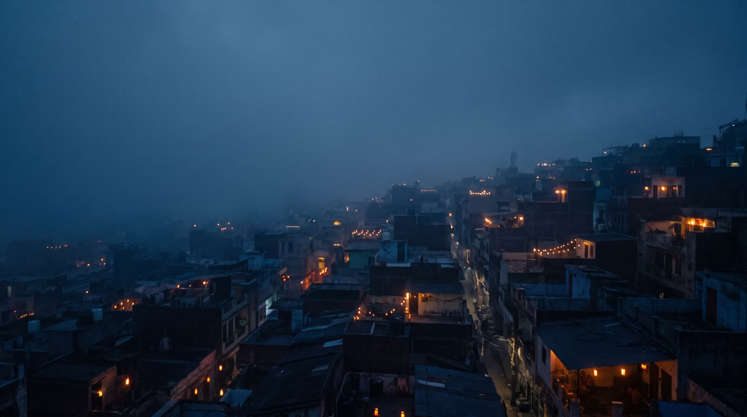 An Indian neighbourhood at blue hour seen from above, warm window and rooftop lights glowing across the dark rooftops